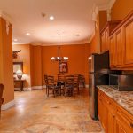 Open kitchen and dining area with wooden cabinets, granite countertop, stainless steel sink, microwave, fridge, and a round dining table with six chairs under a chandelier. Warm orange walls and decorative moldings.