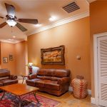 A cozy living room with brown leather sofas, a wooden coffee table, a ceiling fan, Southwestern-style decor, and warm orange walls. An adjoining kitchen with wood cabinets is visible in the background.