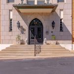 The entrance to a building with tan brick walls, double glass doors, two lion statues on either side, potted plants, wall-mounted lanterns, and large steps leading up from the sidewalk. The address "2306" is visible.