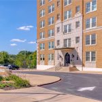 A brick mid-rise apartment building with large windows and a white stone entrance, surrounded by greenery and a paved driveway with a white arrow, on a sunny day with blue sky and clouds.