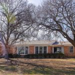 A one-story brick house with white trim surrounded by large leafless trees casting shadows on the front yard under a partly cloudy sky.