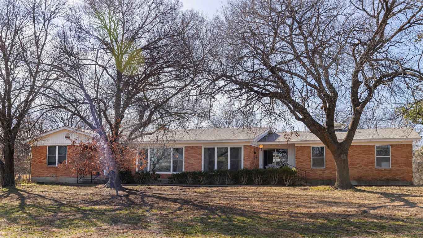 A one-story brick house with white trim surrounded by large leafless trees casting shadows on the front yard under a partly cloudy sky.