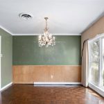 A dining room with green and tan walls, a parquet wood floor, a chandelier, large front windows with beige curtains, and visible outdoor trees through the windows.