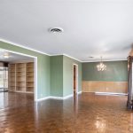Spacious living area with green and tan walls, large windows, wood flooring, a chandelier, built-in shelves, and a ceiling fan. Sunlight streams in from the right, illuminating the open room.