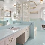 A vintage bathroom with pastel blue tile, a built-in vanity with a pink sink, ornate mirrors, decorative privacy screens, and sheer lace curtains on the windows letting in daylight.
