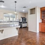A kitchen with light cabinets, granite countertops, a built-in oven, a dishwasher, parquet wood flooring, and a wooden hutch with glass doors. Pendant lights hang over the counter and a large window is above the sink.