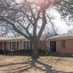 A single-story brick house with large windows and a small front porch, shaded by several leafless trees. Sunlight filters through the branches onto the dry, patchy yard in front.
