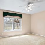 A bright, empty room with beige walls and carpet, a ceiling fan with lights, a large window with a green valance, and a white sliding closet door. Sunlight streams in through the window.