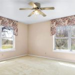 Empty room with beige walls and carpet, two large windows with floral valances, and a ceiling fan with lights. Sunlight streams in, and trees are visible outside the windows.
