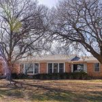 A one-story brick house with large windows sits behind leafless trees on a sunny day. The yard in front is mostly bare, with patches of grass and fallen leaves scattered on the ground.