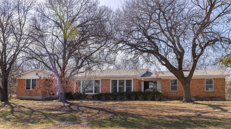 A one-story brick house with large windows sits behind leafless trees on a sunny day. The yard in front is mostly bare, with patches of grass and fallen leaves scattered on the ground.