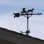 A weather vane shaped like a horse pulling a carriage sits atop a roof, silhouetted against a cloudy sky.