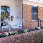 A small brick patio outside a house features a white metal table and chairs, a potted plant on the table, and a flower bed with colorful flowers along a low brick wall. A large window and sliding door are visible.