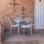 A small patio with a light blue metal table and four matching chairs. A pot of flowers sits on the table. The brick wall behind features two metal crosses, and a round wall light is mounted nearby.