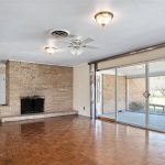 Spacious living room with wood parquet flooring, a brick fireplace, ceiling fans, a grandfather clock, and large sliding glass doors leading to a bright sunroom with outdoor views.