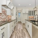 Modern kitchen with white cabinets, brick backsplash, black countertops, stainless steel appliances, large island with sink, pendant lights, and light wood flooring. Bright and spacious with an open layout.