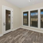 A bright, empty room with light beige walls, wood-style flooring, a glass door, and three large windows with dark blinds, overlooking a grassy yard.