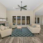Modern living room with beige sofas, armchairs, a patterned blue rug, a brick fireplace, built-in shelves, ceiling fan, and open kitchen area with pendant lighting and a breakfast bar. Large windows along the back wall.