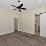 A modern empty bedroom with beige walls, dark carpet, a black ceiling fan, and a white sliding barn door leading to a bathroom. Large windows on the right let in natural light.