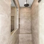 Modern walk-in shower with beige stone tiles, a built-in bench, a rainfall showerhead on the ceiling, a black wall-mounted handle, and a glass door; features a large window into an adjacent room.