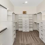 A spacious walk-in closet with white built-in shelves, drawers, and hanging rods on both sides, plus carpeted flooring and a small box on the top shelf at the far end.