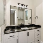 Modern bathroom with a glass shower, black countertop, white vanity cabinets, rectangular mirror, black fixtures, and wall-mounted light fixture. A window with blinds lets in natural light above the sink.