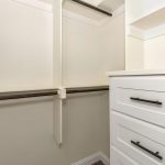 Empty walk-in closet with white walls, built-in white drawers with black handles, and multiple dark metal hanging rods. An electrical outlet is visible above the drawers.