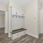 A modern entryway with light beige walls, white doors, wood-style flooring, and a built-in bench with storage cabinets and coat hooks. Multiple doorways lead to other rooms in the house.