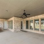 A covered patio area with a ceiling fan, recessed lighting, and brick walls. There are multiple windows with blinds, a door, and a built-in counter on the left side. The floor is concrete.