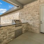 Covered patio area with a built-in stainless steel outdoor grill and storage cabinets set in a brick counter, next to a white door; view of a grassy yard and trees under a blue sky with clouds.