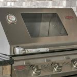 A stainless steel outdoor grill with a glass window on the lid, a built-in thermometer, and three control knobs, set into a stone countertop with a brick wall in the background.