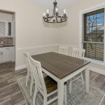 A dining room with a wooden table, four chairs, light-colored walls, wood flooring, and a large window. A chandelier hangs above the table, and a built-in cabinet with shelves is visible in a nook.