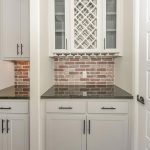 White kitchen cabinets with black countertops, a brick backsplash, and glass-front upper cabinets with a built-in wine rack. Stainless steel appliances are visible on the left, and a white door is on the right.