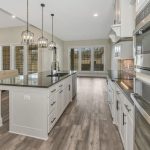 Modern kitchen with light gray cabinets, stainless steel appliances, a large island with a dark countertop, pendant lights, and wood-look flooring. An open living area with a fireplace and large windows is visible in the background.