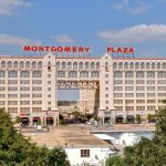 A large historic building with “Montgomery Plaza” in red letters on top, featuring white arches and many windows. Trees and smaller buildings are in the foreground under a blue sky with scattered clouds.