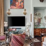 Eclectic living room with patterned chairs, a glass table, animal print rug, ornate mirror, and a TV below a colorful portrait. Large window with gold curtains lets in natural light.