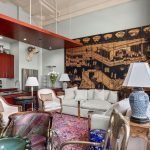 A stylish living room with a kitchenette, a large wall tapestry, eclectic furniture, patterned rug, decorative lamps, and tall windows letting in natural light.