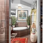 A modern bathroom with brown tiled walls, a corner bathtub, potted plants, framed artwork above the tub, a red patterned rug, and a glass shower door partially open.