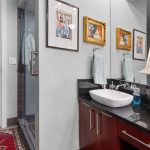 A modern bathroom with a black countertop, white sink, floral lamp, and framed art on the wall. A glass shower is to the left, and a bedroom and closet are visible through open doors in the background.