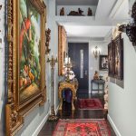 A hallway with dark wood floors and a vibrant red patterned rug, featuring ornate framed paintings, antique furniture, and decorative statues, leading to a door at the end.