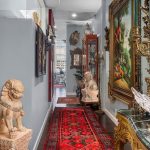 A hallway with dark wood floors features a red Persian rug, ornate framed paintings, decorative statues, and antique furniture, leading toward large windows with a city view.