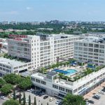 Aerial view of Montgomery Plaza, a large white multi-story building complex with a rooftop pool and surrounding cityscape, trees, and parking lots under a clear blue sky.