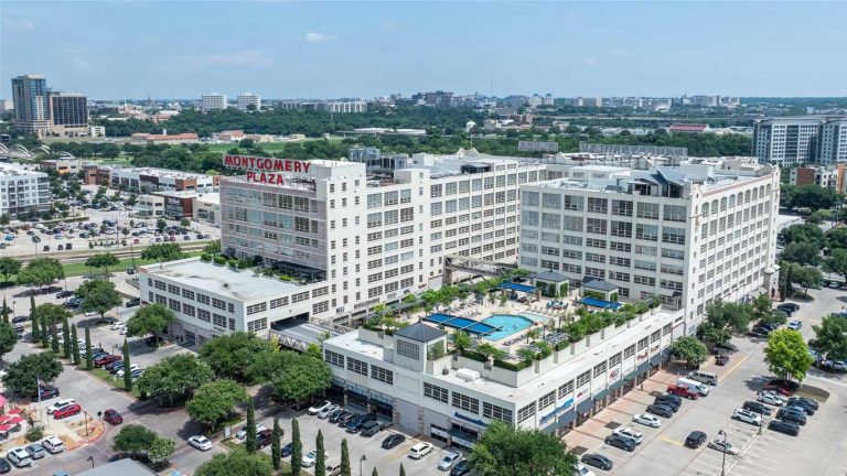 Aerial view of Montgomery Plaza, a large white multi-story building complex with a rooftop pool and surrounding cityscape, trees, and parking lots under a clear blue sky.