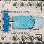 Aerial view of a rectangular outdoor swimming pool with people swimming, surrounded by lounge chairs, sunshades, greenery, and parked cars along the left edge.