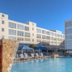 A large outdoor swimming pool with lounge chairs, umbrellas, and cabanas, set beside modern white multi-story buildings under a clear blue sky. Stone walls frame part of the pool area.
