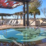 Outdoor hot tub with clear water, steps, and metal handrails, surrounded by lounge chairs and shaded seating areas. Trees and open sky are visible in the background on a sunny day. Signs read "NO DIVING.