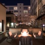 Outdoor seating area at night with four cushioned chairs arranged around a lit fire pit, surrounded by modern buildings with large windows and minimal landscaping.