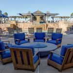 Outdoor lounge area with six blue cushioned chairs around a fire pit, facing a stone water feature. Lounge chairs, umbrellas, and trimmed bushes are visible in the background under a clear sky.