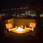 Four cushioned chairs surround a fire pit on a rooftop patio at night, with city lights and buildings visible in the background.
