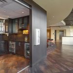 Modern office kitchen and lounge area with dark cabinets, glass shelves, under-counter fridge, and bar seating by large windows. Curved hallway leads to open space with more seating and natural light.